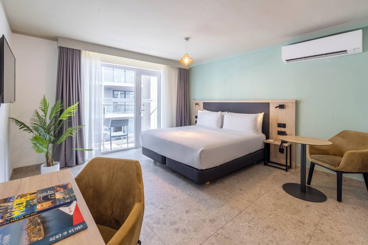 A hotel room with a bed, a desk, a chair, a potted plant, and a view of the outside balcony.