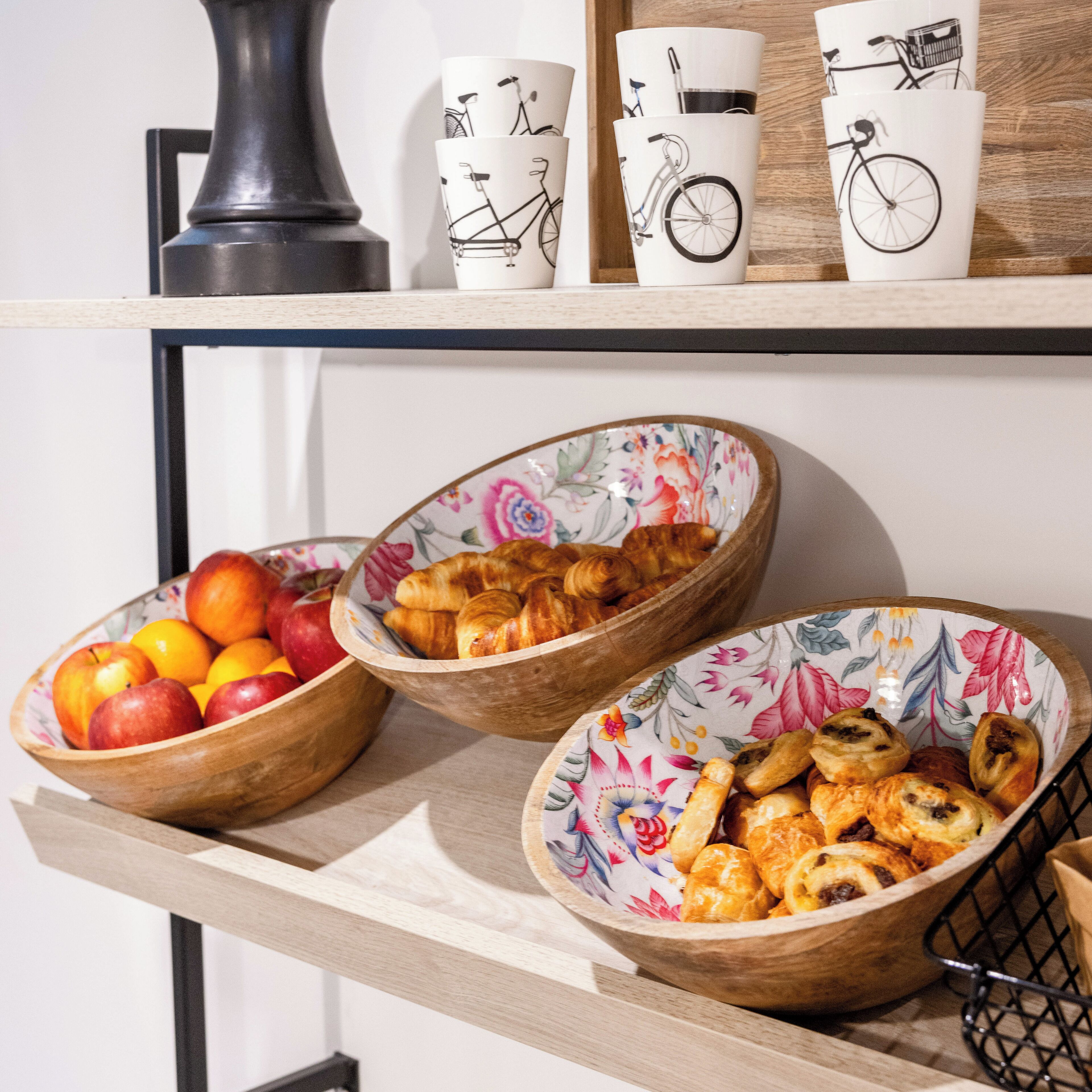 Una estantería con tres cuencos de madera que contienen frutas y pasteles, junto con tres tazas blancas decoradas con diseños de bicicleta.
