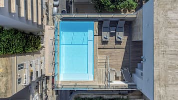An aerial view of a rooftop pool with a clear blue pool, surrounded by a wooden deck and lounge chairs.