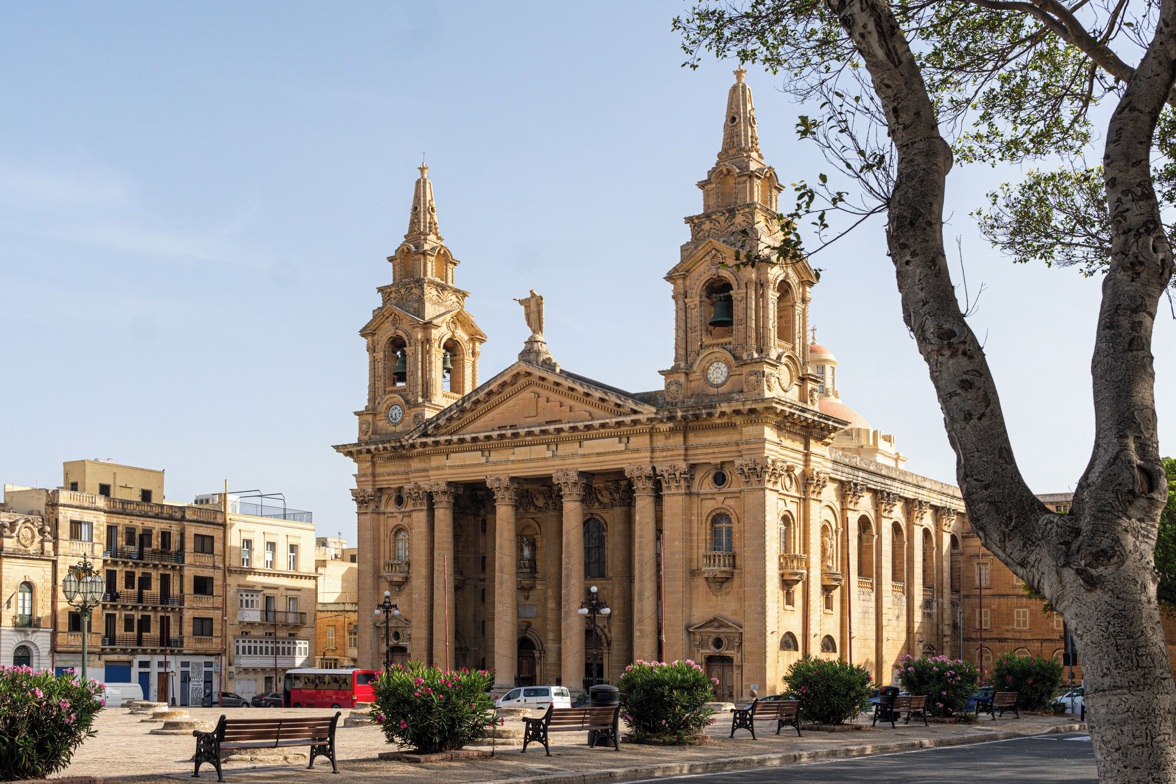 A historic building with two prominent towers and a central dome, surrounded by benches and greenery.