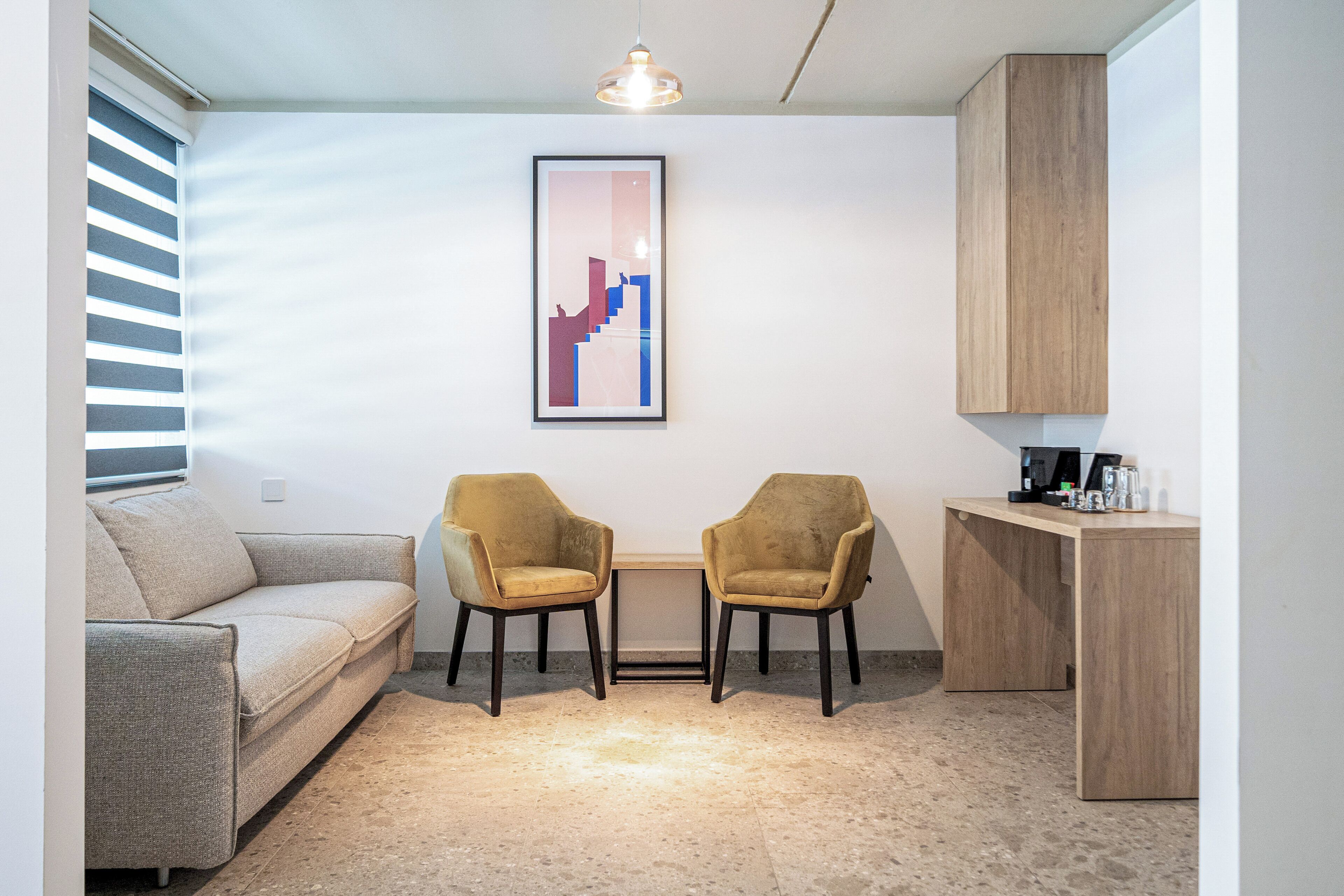 A modern room with a sofa, two chairs, a small table, a framed artwork, a cabinet, and a coffee machine.