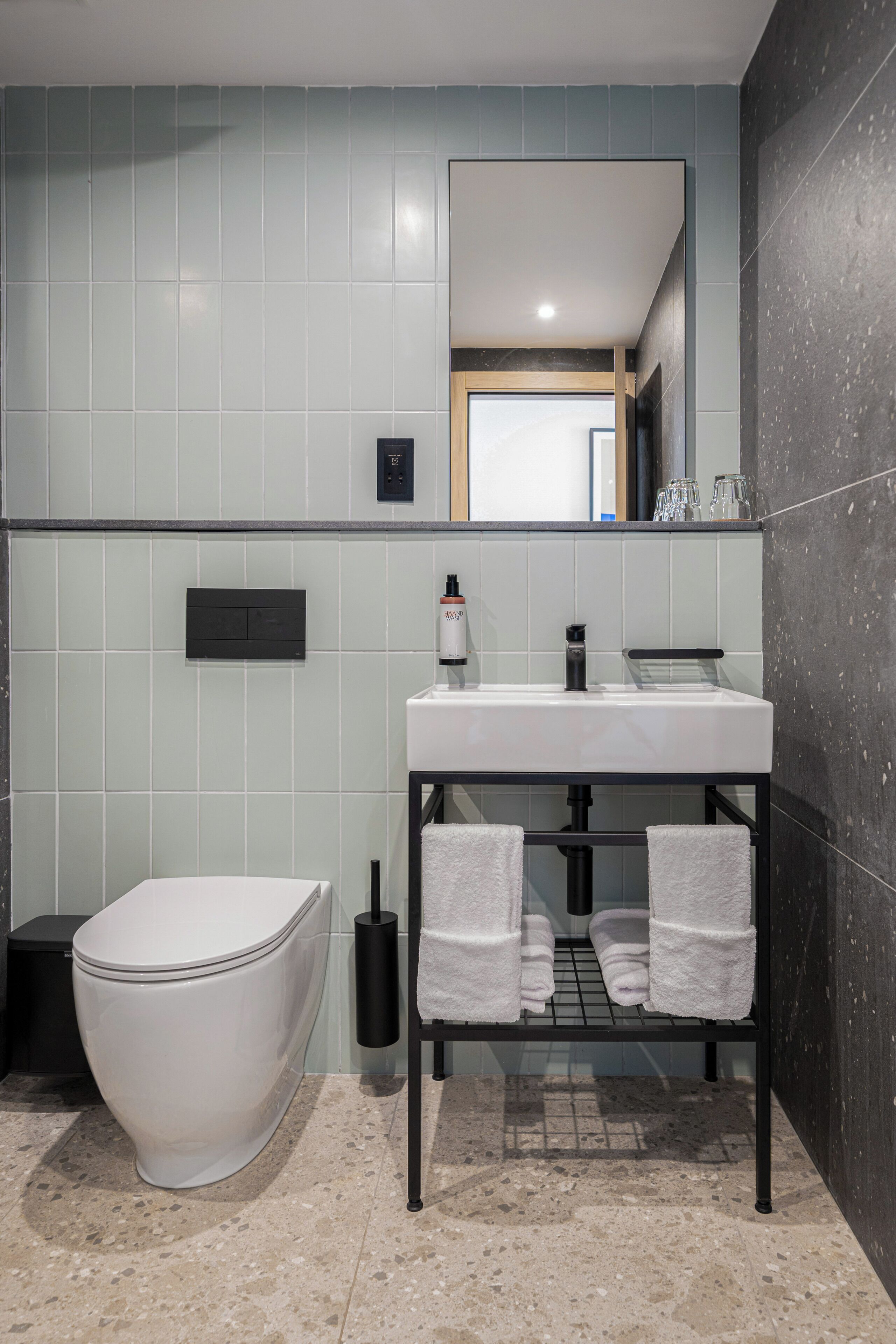Un baño moderno con inodoro blanco, lavamanos con encimera negra y espejo sobre el lavamanos. Hay toallas colgadas en un perchero debajo del lavamanos.