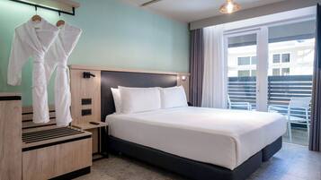 A hotel room with a large bed, a wooden nightstand, a robe rack with two robes, and a view of the outside balcony.