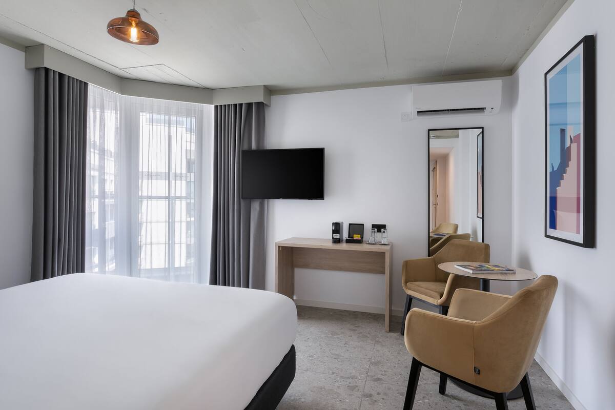 A hotel room with a large bed, a desk with a flat-screen TV, a chair, and a small table.