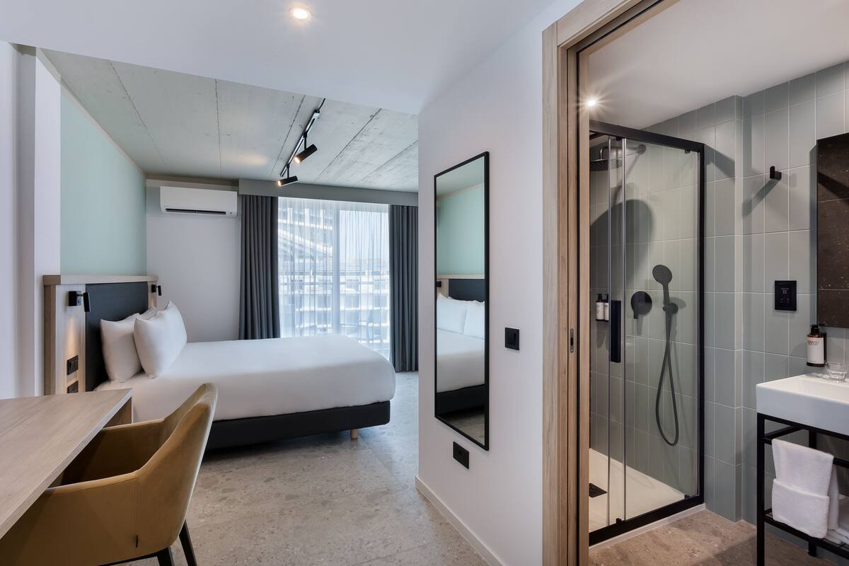 A hotel room with a bed, a desk, a chair, a shower, and a bathroom with a sink.