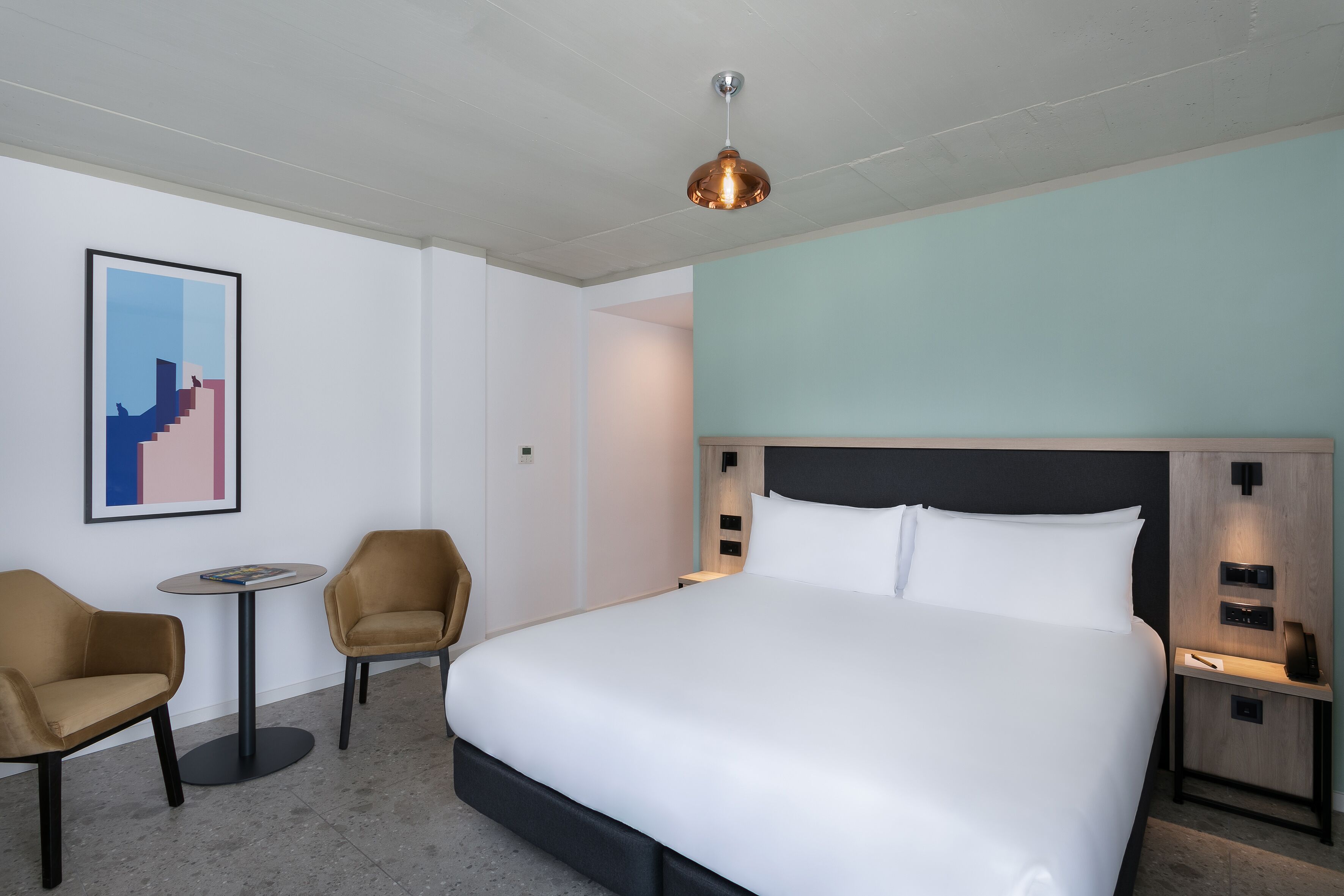 A hotel room with a large bed, a small round table, two chairs, a wall-mounted artwork, and a modern pendant light.