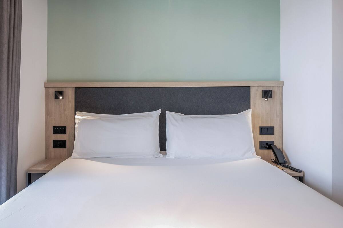 A neatly made bed with white linens, a wooden headboard, and a bedside table with a phone and a power outlet.