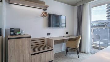 A modern hotel room with a wooden desk, a chair, a flat-screen TV mounted on the wall, and a balcony with a view of the city.