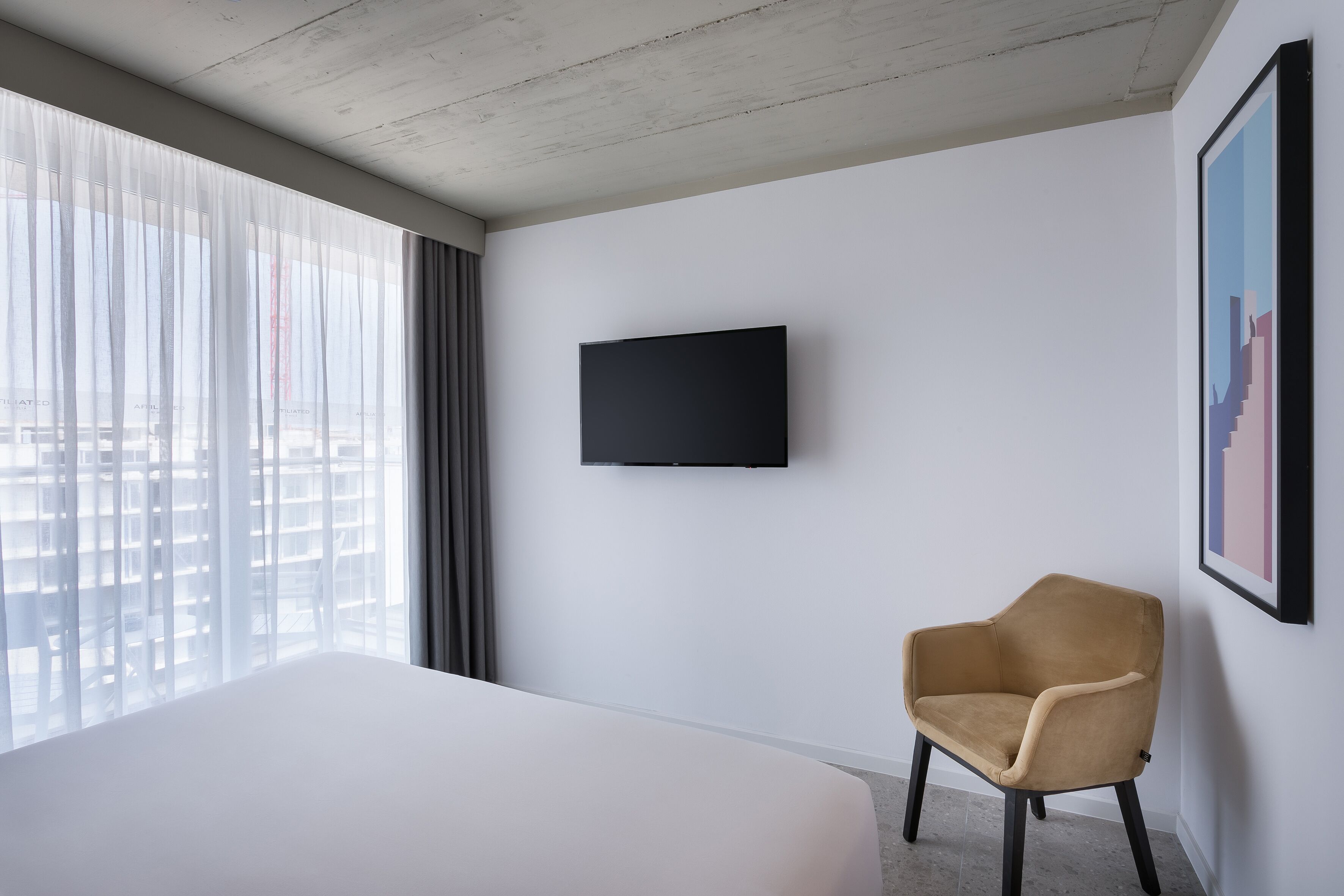 Habitación de hotel con una cama grande, un televisor montado, una silla y vistas a la ciudad a través de cortinas translúcidas.