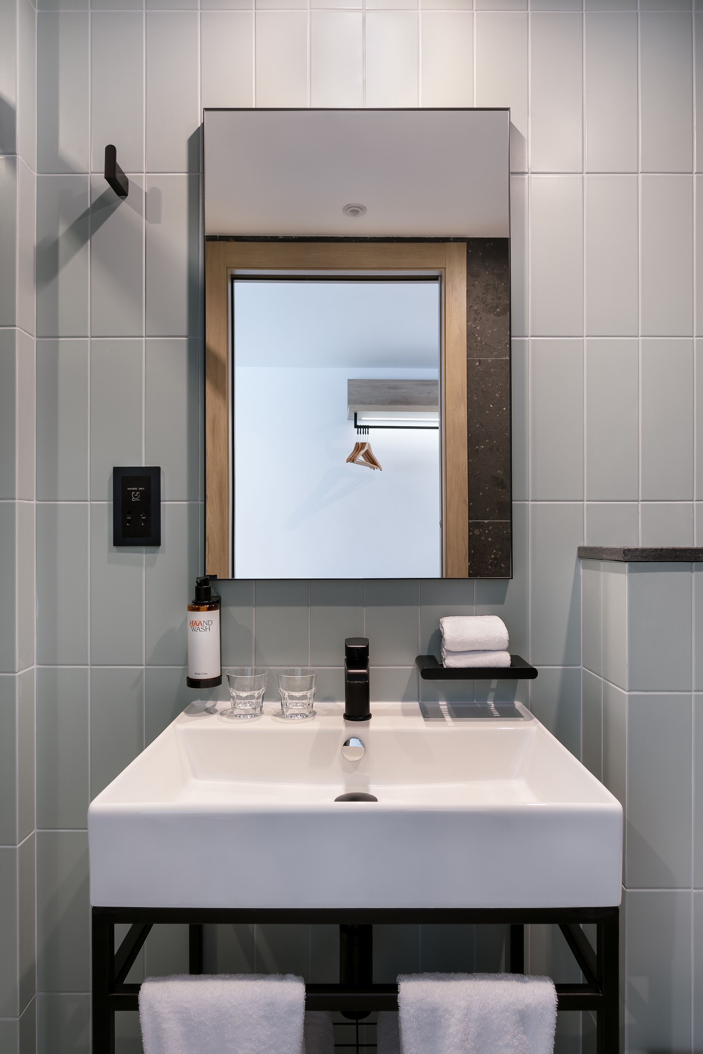 A modern bathroom with a white sink, a mirror with a wooden frame, a towel rack, and a hand wash dispenser.