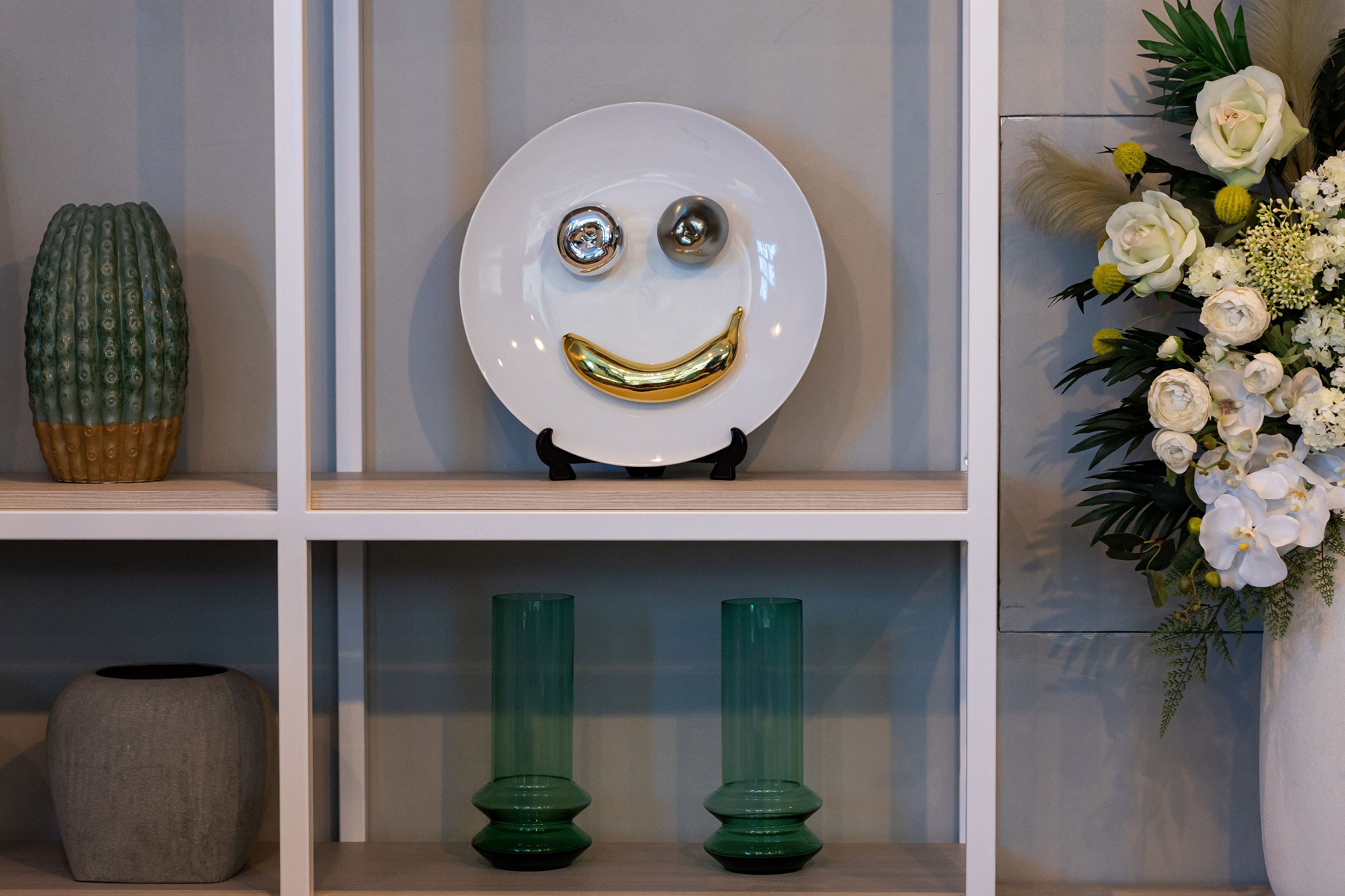 Uma prateleira com um cacto, um prato sorridente e dois vasos verdes.