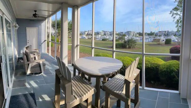 Un patio techado con una mesa y sillas, y amplios ventanales que ofrecen vistas a un puerto deportivo y edificios.