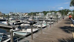 Un puerto deportivo con numerosas embarcaciones amarradas, un paseo marítimo de madera y casas al fondo.