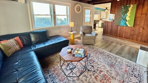 A cozy living room with a black leather sofa, a wooden coffee table with books, a patterned rug, and a view of a kitchen and dining area through an open door. - Sand Life - Pelican's Northern Nest -Pet Friendly! (Kill Devil Hills)