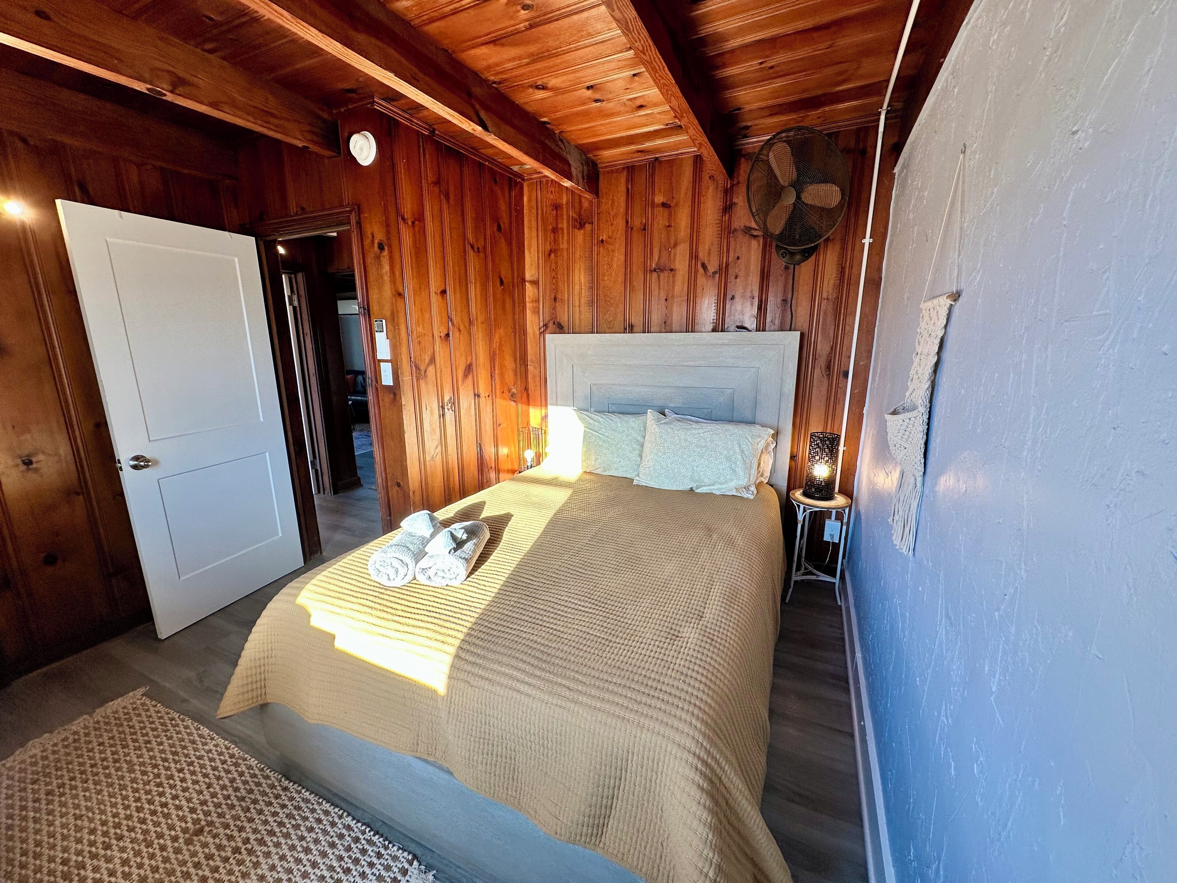 A wooden-paneled bedroom with a bed, a nightstand, and a wall-mounted fan.