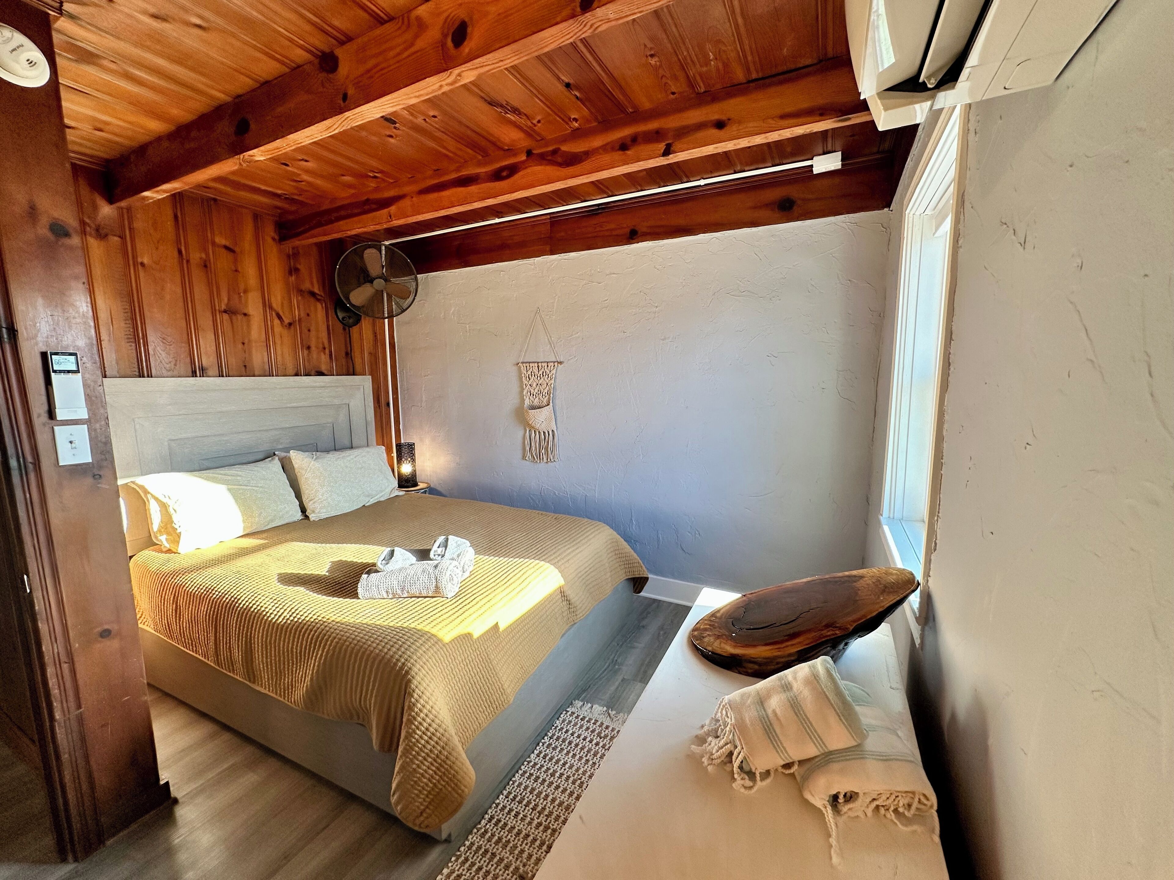 Une chambre confortable avec un plafond en bois, un lit recouvert d’une couverture beige, une table de chevet avec une lampe et une décoration murale en macramé.