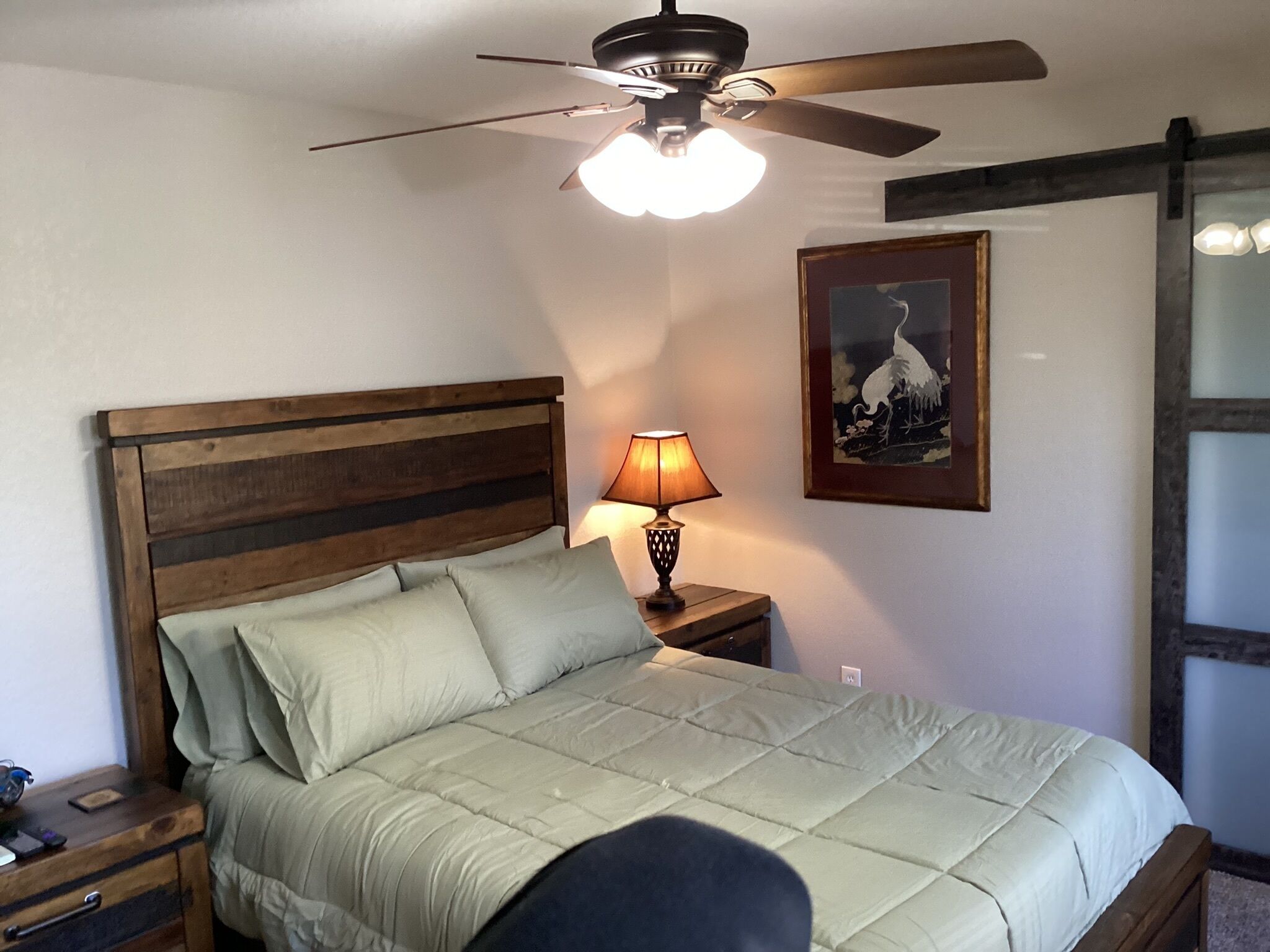 A bedroom with a wooden headboard, a bed with a light-colored quilt, a bedside lamp, and a ceiling fan.