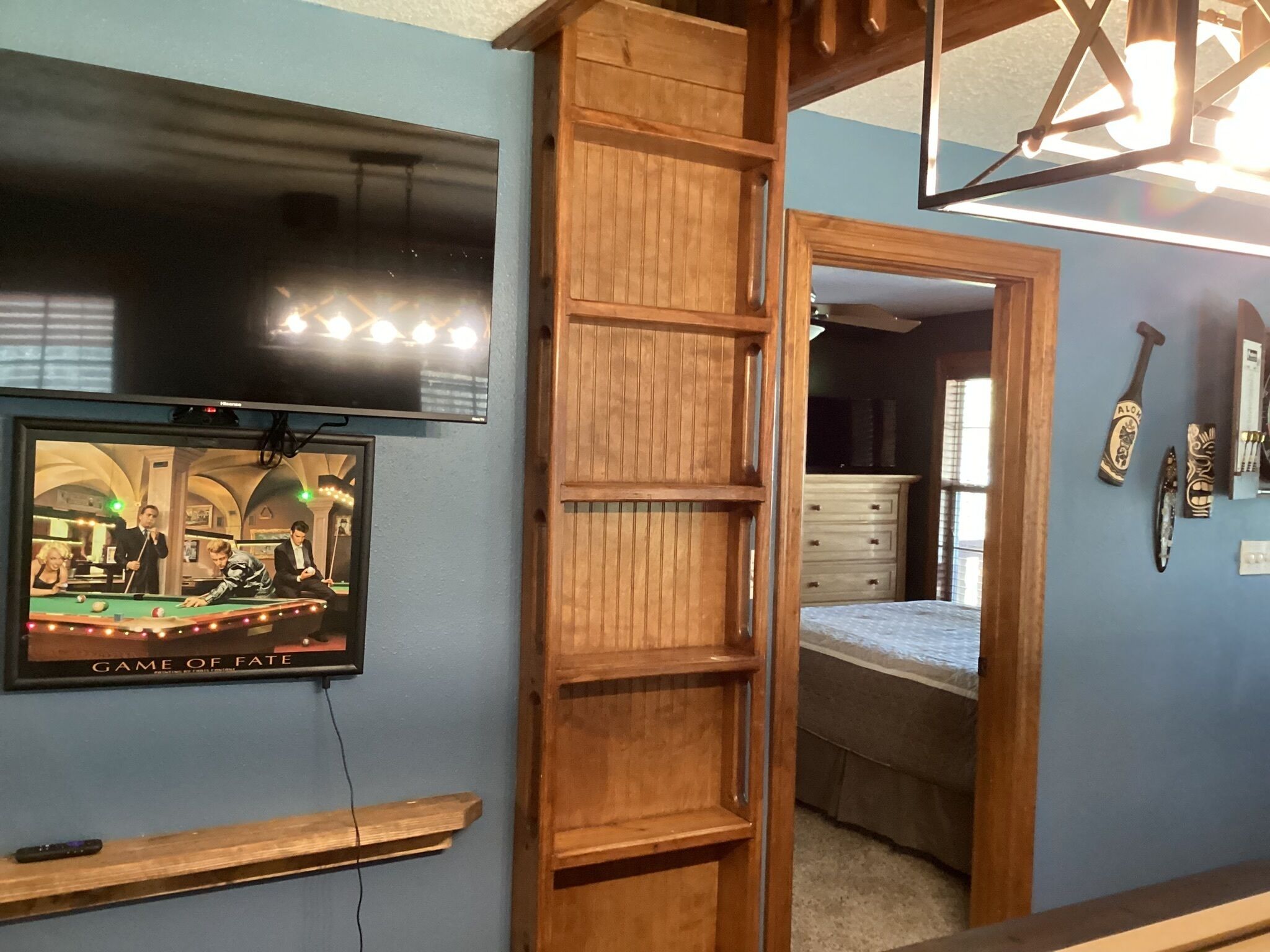 Una habitación con una televisión montada en la pared, un estante de madera, una cama y un aparador.