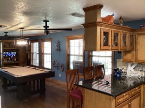 Relax in this spacious living room with a pool table, comfortable seating, and a kitchen area, featuring a bar with a neon sign.