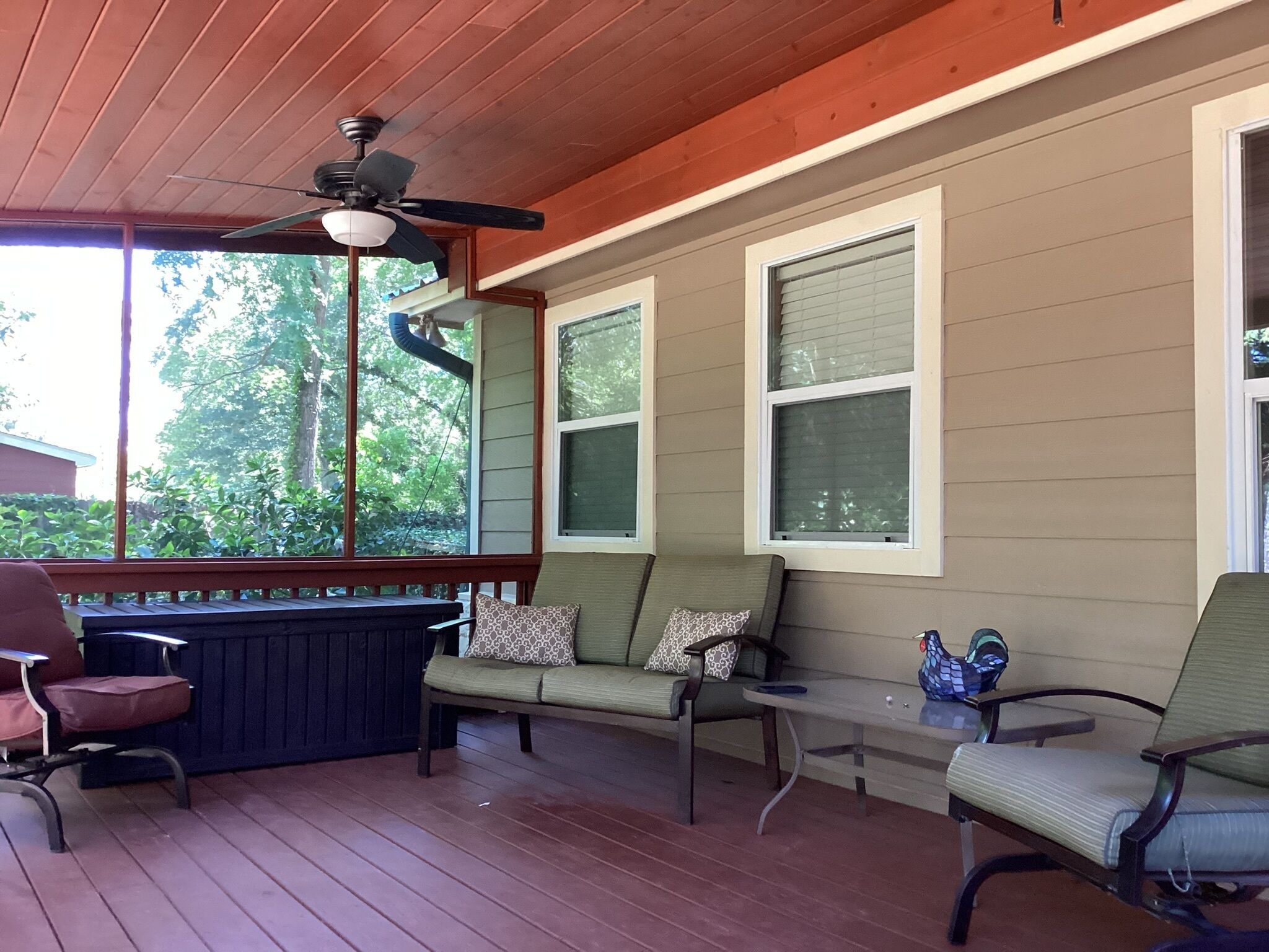 A covered porch with a wooden ceiling, a ceiling fan, a green sofa with patterned cushions, a small table, and a chair.