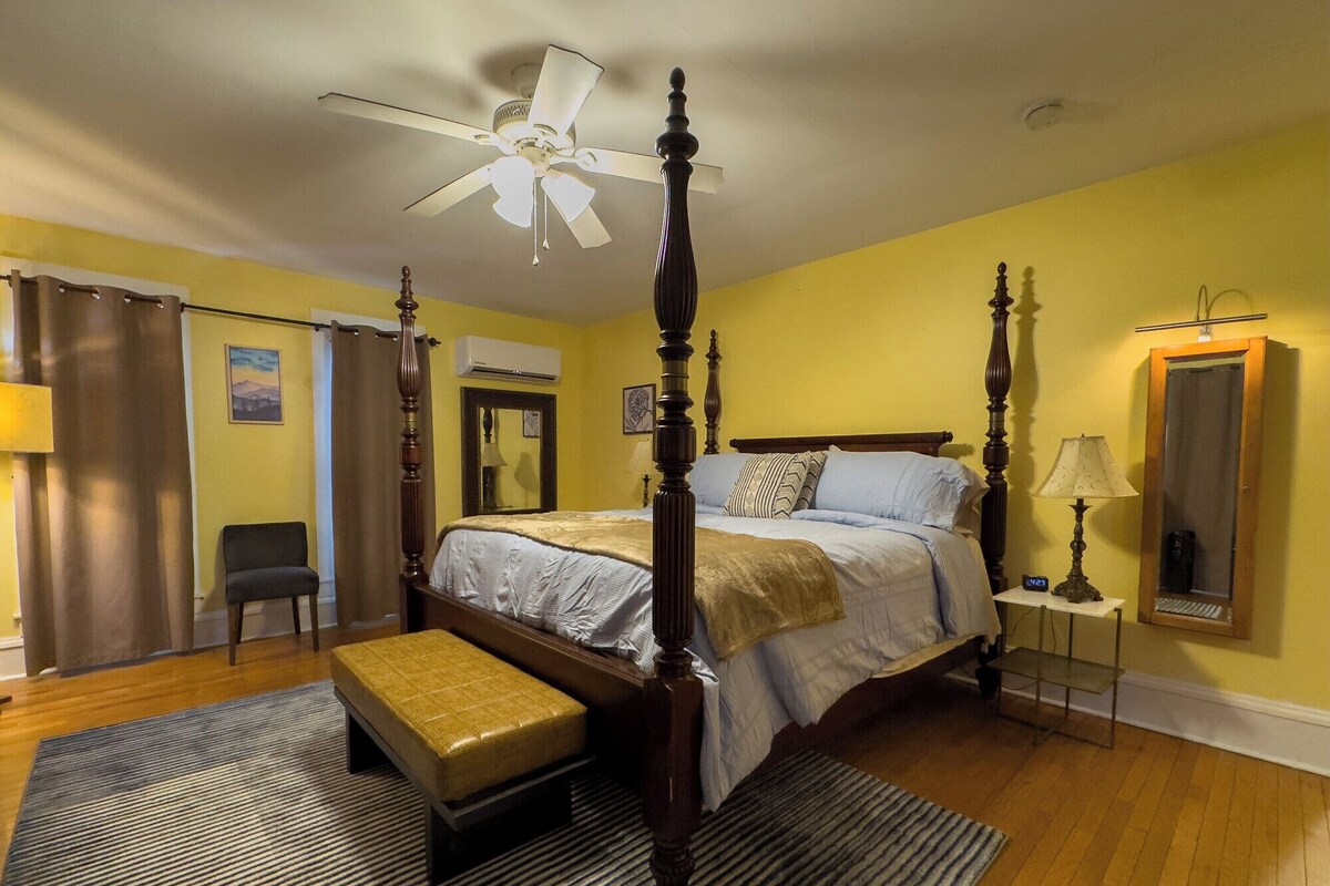 A four-poster bed with a canopy, a bedside table with a lamp, a chair, and a bench in a room with yellow walls and wooden flooring.