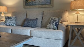 A living room with a grey sofa, patterned cushions, a coffee table, a painting of boats, and two lamps. - Redesigned condo in oceanfront community! (St. Simons Island)