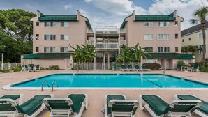 A swimming pool with lounge chairs, a building with balconies, and palm trees. - Redesigned condo in oceanfront community! (St. Simons Island)