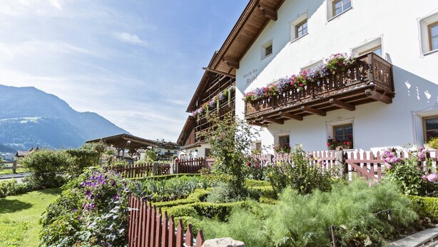 Ein traditionelles, im alpinen Stil gehaltenes Gebäude mit hölzernen Balkonen, die mit Blumen geschmückt sind, umgeben von gepflegten Gärten und einer Bergkulisse.