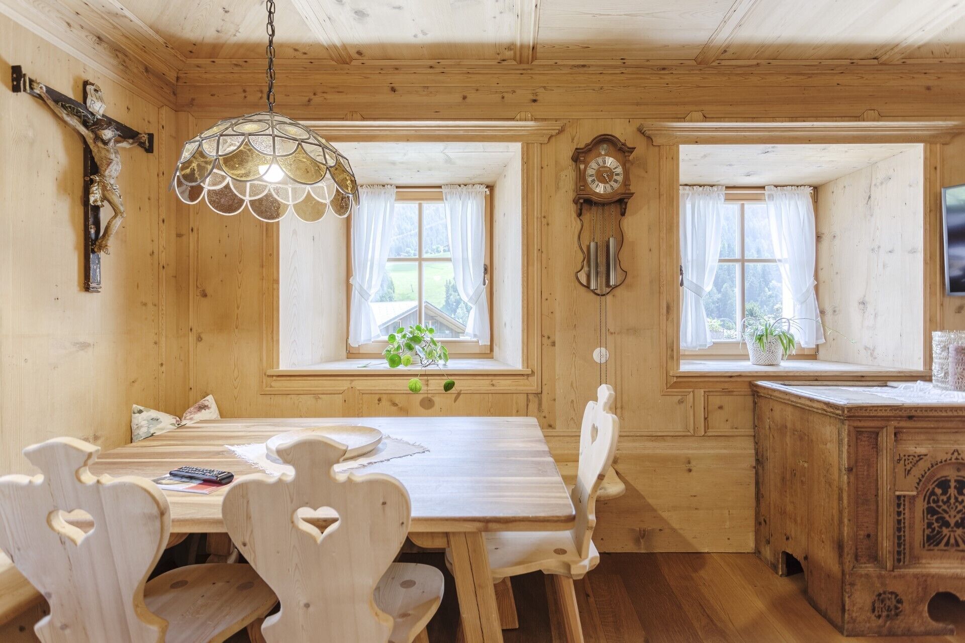 Un intérieur en bois comprenant une table à manger, des chaises, un luminaire suspendu, une horloge murale et des fenêtres avec des rideaux.