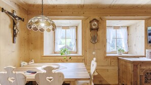Ein Holzinterieur mit einem Esstisch, Stühlen, einer Pendelleuchte, einer Wanduhr und Fenstern mit Vorhängen.