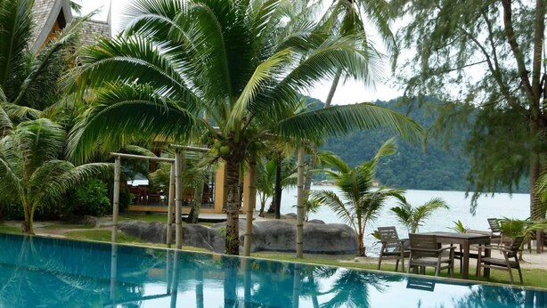 A pool area with a view of a lake, surrounded by palm trees and a wooden deck with chairs. - Villa Ban Rim Nam 50c (Ko Chang)