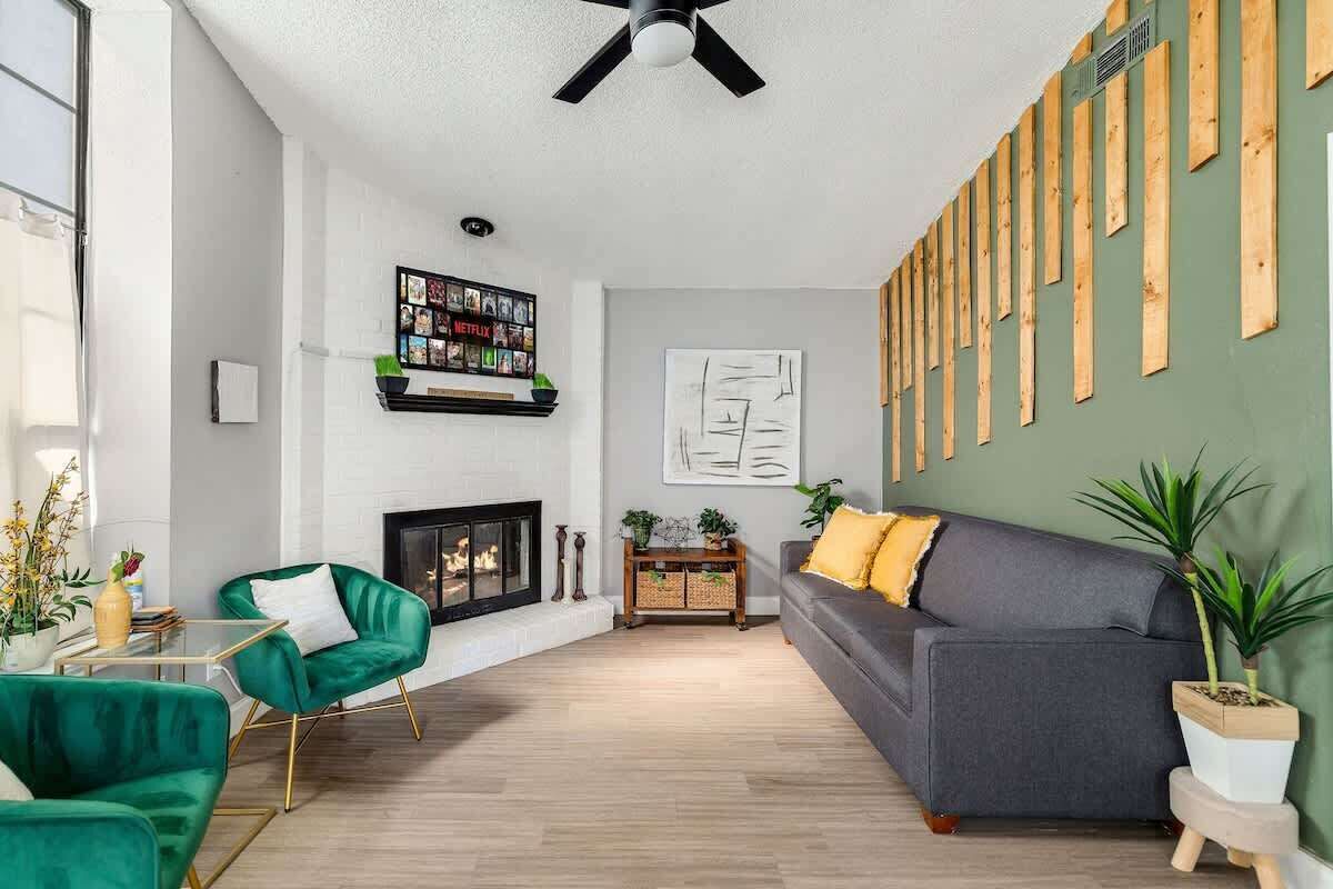 Ein modernes Wohnzimmer mit Kamin, grünen und grauen Sofas, Deckenventilator und einer an der Wand befestigten Holzverkleidung.