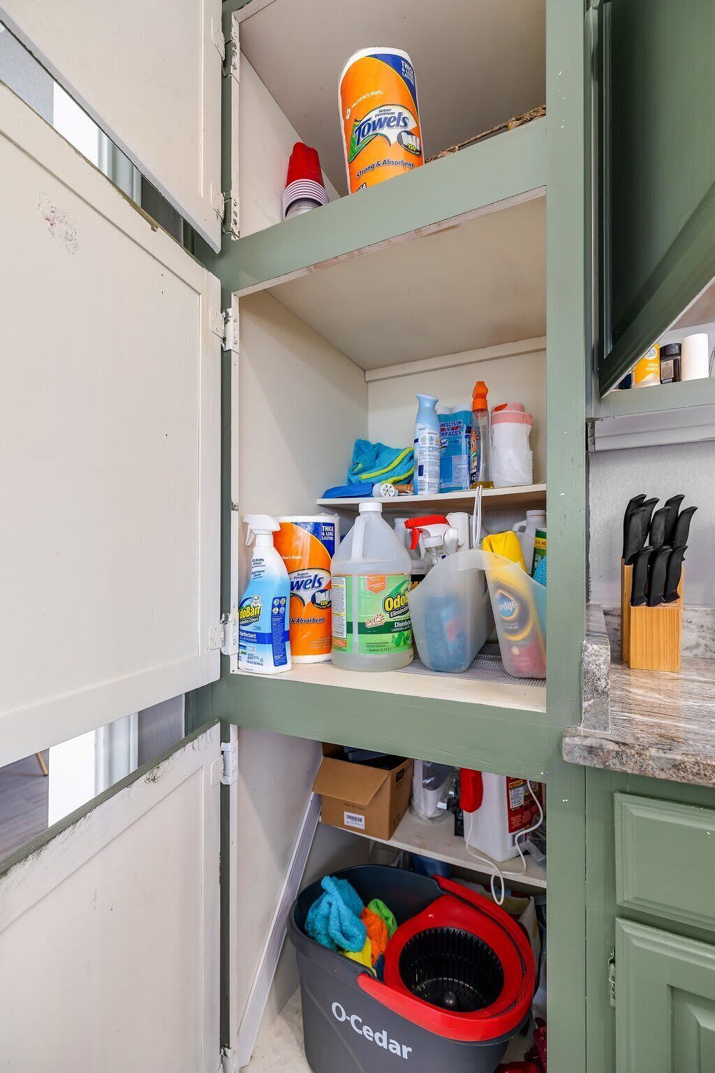 A kitchen with a shelf containing cleaning supplies, a trash bin, and a knife block.