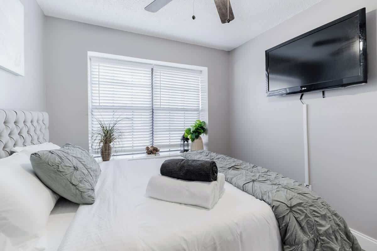 A bedroom with a bed, a TV mounted on the wall, and a window with blinds.