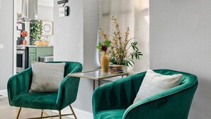 Un salon moderne avec deux fauteuils en velours vert, une petite table, une fenêtre donnant sur un beau paysage et une cuisine en arrière-plan.