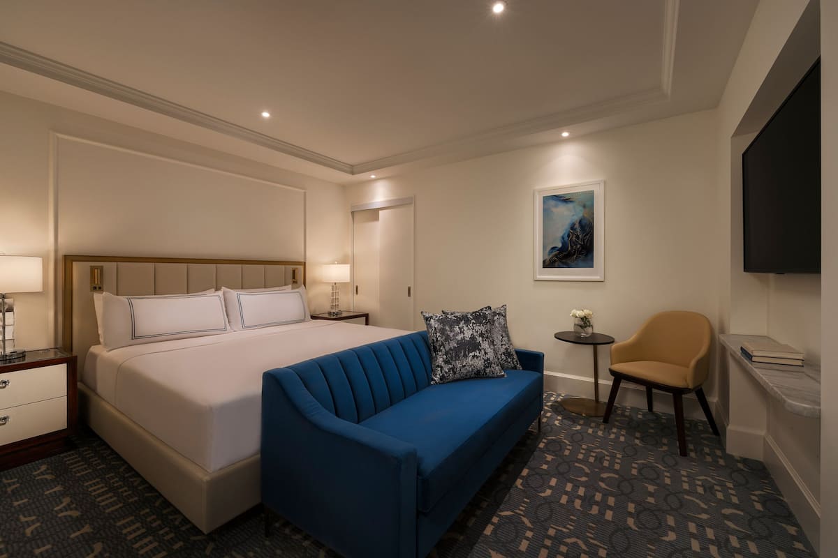 A hotel room with a large bed, a blue sofa, a small table with a vase, and a TV mounted on the wall.