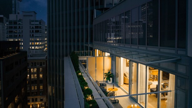 Un edificio moderno con fachada de vidrio, un balcón con mobiliario exterior y vistas de la ciudad iluminada de noche.