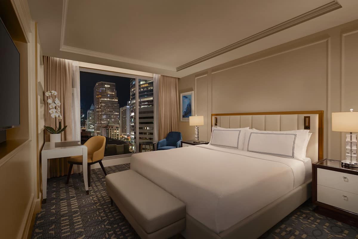 A hotel room with a large bed, a desk, a chair, a nightstand, and a view of a cityscape at night.