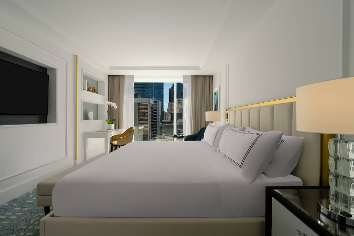 A modern hotel room with a large bed, a television, a desk with a chair, and a view of the cityscape through large windows.