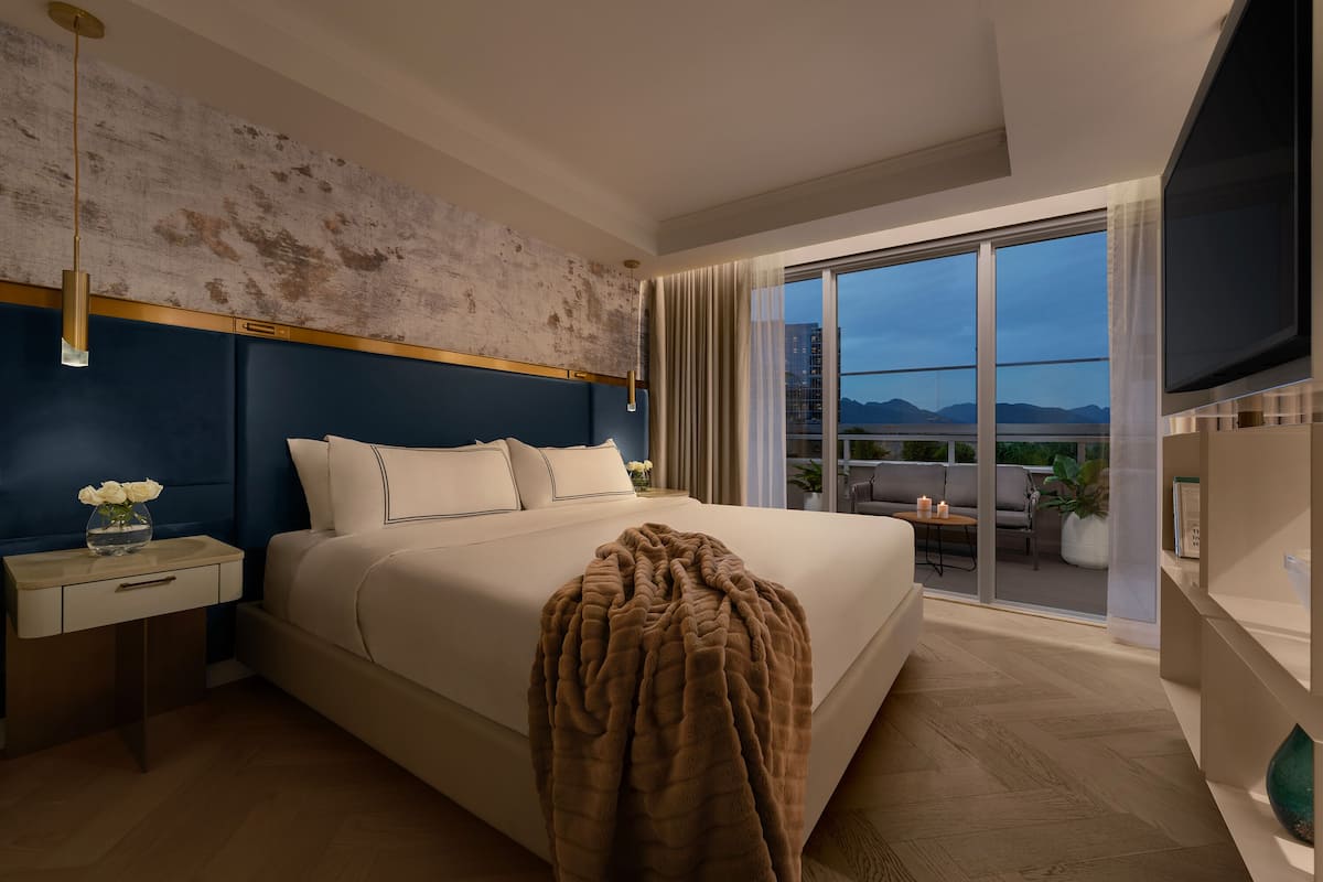A modern hotel room with a large bed, a nightstand, a TV, and a balcony with a view of the mountains.