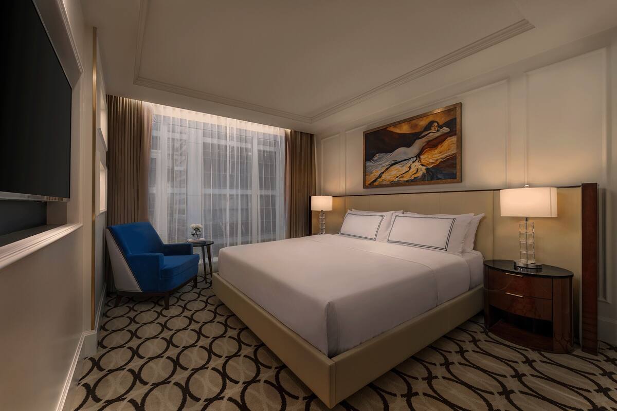A hotel room with a large bed, a blue armchair, a small table, and a painting on the wall.