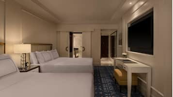Quarto de hotel com duas camas, uma mesa, uma televisão e um banheiro visível através de uma porta aberta.