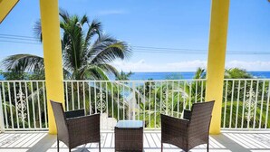 Ein Balkon mit zwei Korbstühlen und einem kleinen Tisch, der Blick auf einen Strand mit Palmen und das Meer freigibt.