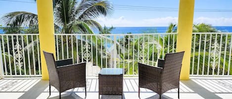 A balcony with two wicker chairs and a small table, overlooking a beach with palm trees and the ocean.