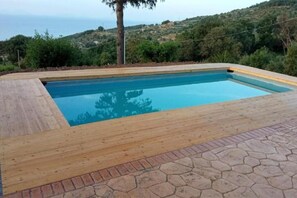 Une piscine rectangulaire dotée d’une terrasse en bois et bordée de pierres, entourée de verdure et avec des collines en arrière-plan.