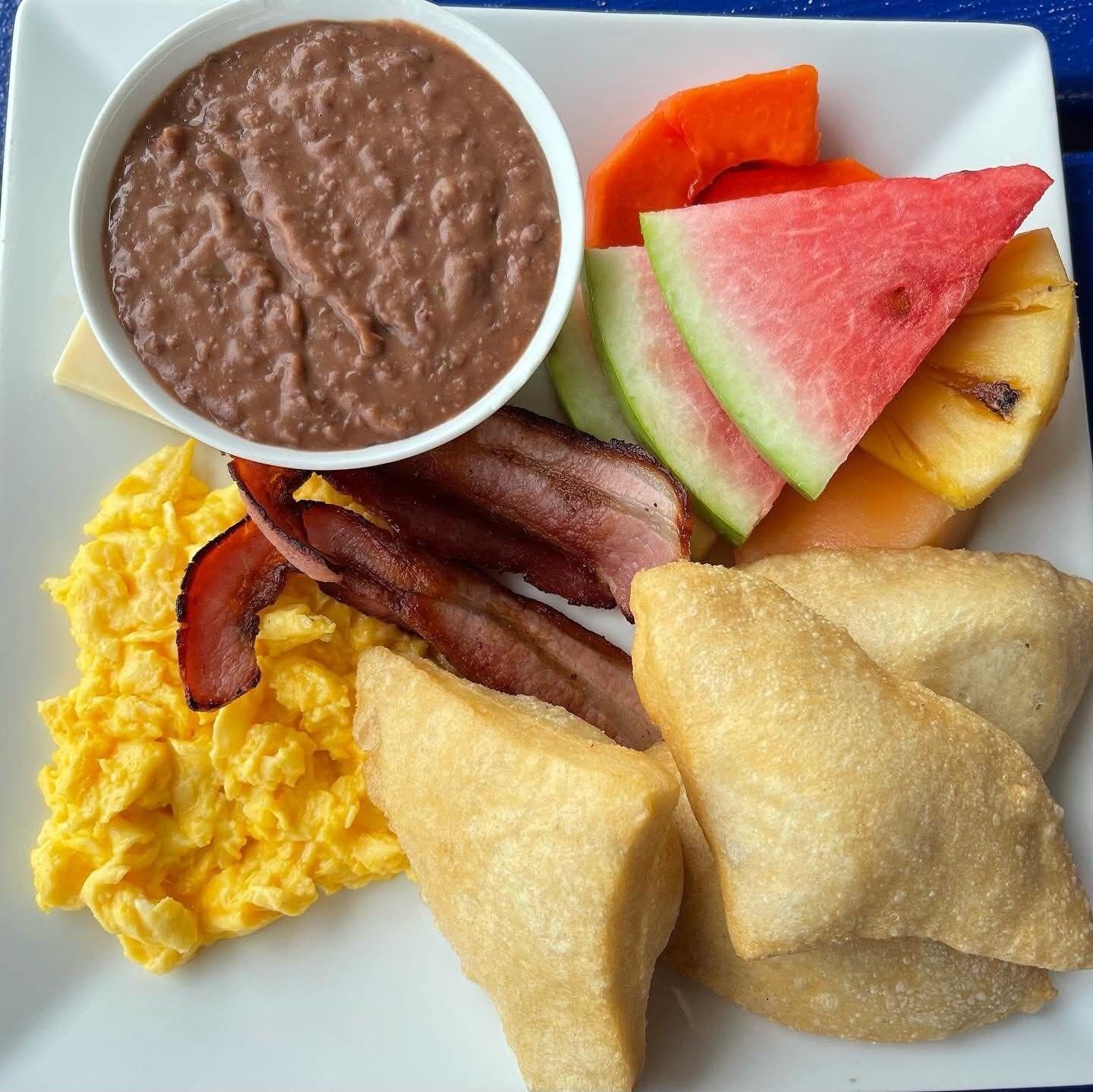 Un plato con huevos revueltos, tocino, bollería frita, un tazón de frijoles y rebanadas de sandía.