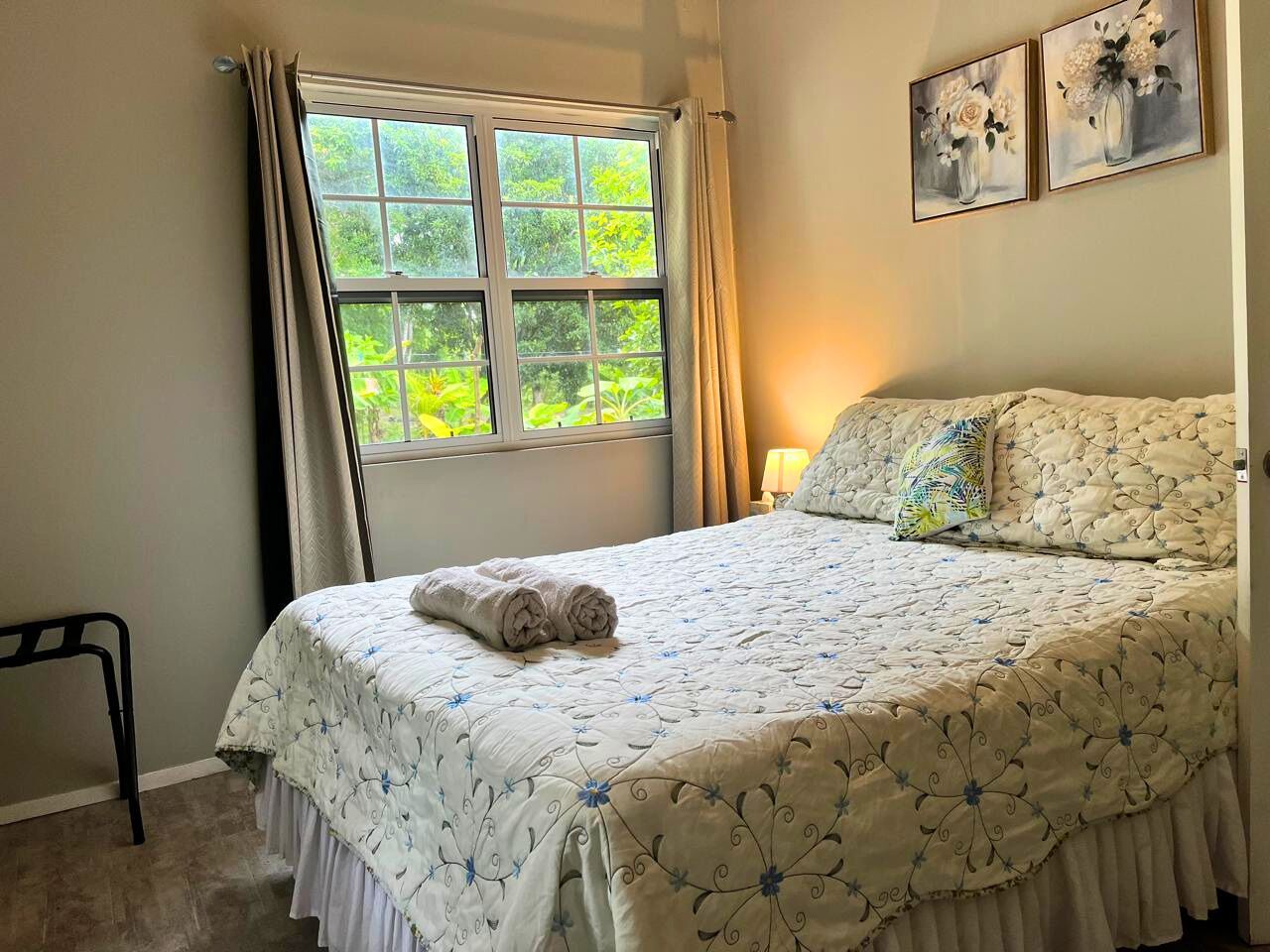 Una cama bien tendida con una colcha estampada, una toalla enrollada al lado y una ventana con cortinas.