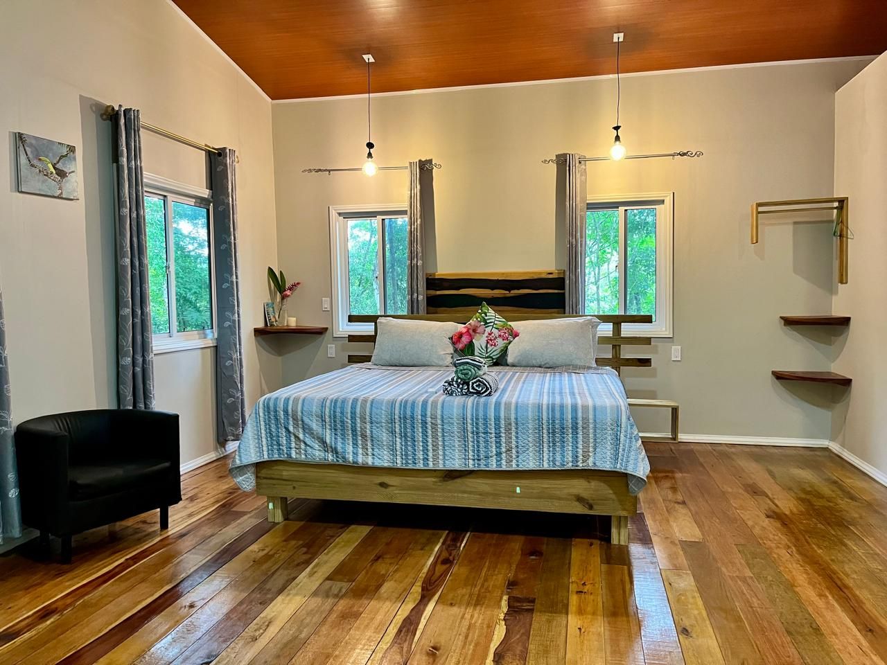 A bedroom with a wooden bed, two windows with curtains, a black chair, and a wooden floor.