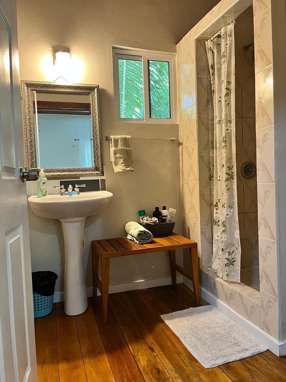 Une salle de bain avec un lavabo sur pied, un miroir, une fenêtre, une douche avec des rideaux à motifs floraux et une petite table en bois sur laquelle sont posés des articles de toilette.