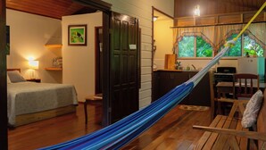A room with a bed, a hammock, a small table, a chair, a kitchen area with a stove, and a window with curtains. - The Cabins at Monkey Fall (San Ignacio)